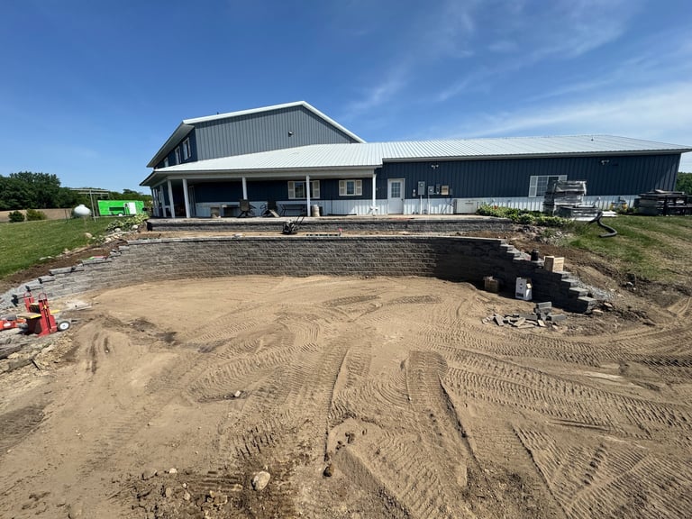 Modern black barn with white trim during construction, showing excavated dirt foundation area in foreground under blue sky