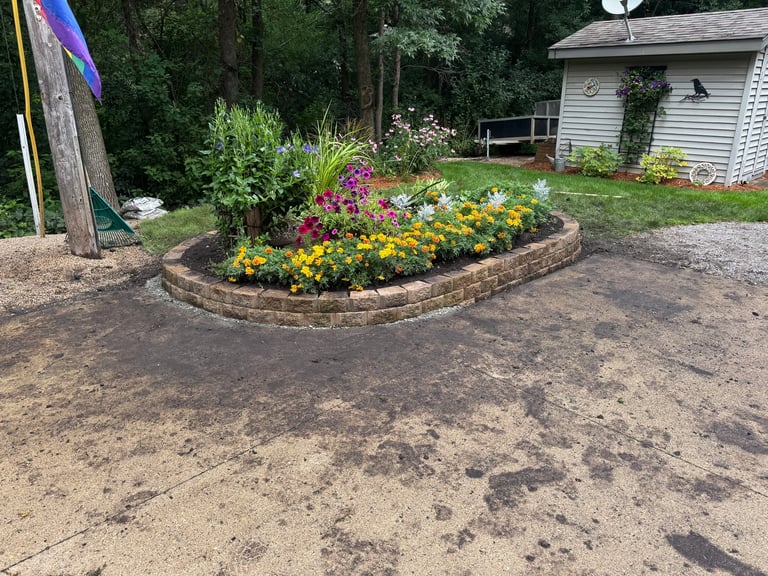 A brick-lined garden bed with colorful flowers including pink, yellow, and purple blooms in a residential yard with a house and trees in the background.