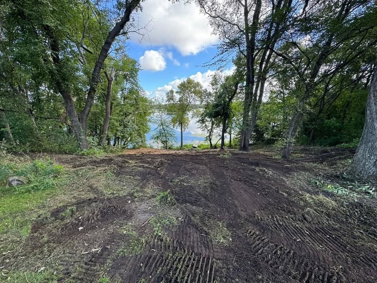 Freshly tilled soil on park grounds lined with mature trees, blue sky with clouds visible between canopy