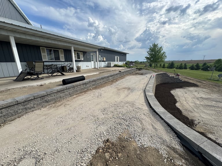 Modern farm building with white trim and covered porch, adjacent to gravel driveway under construction with curved black edging against rural landscape and cloudy sky