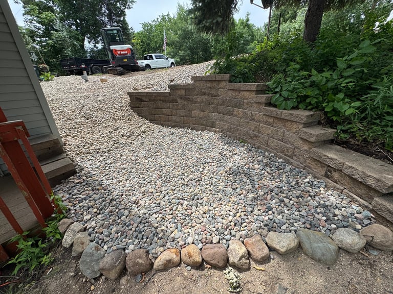 Gravel driveway with curved retaining wall, decorative stones, parked vehicles, and surrounding trees and landscaping