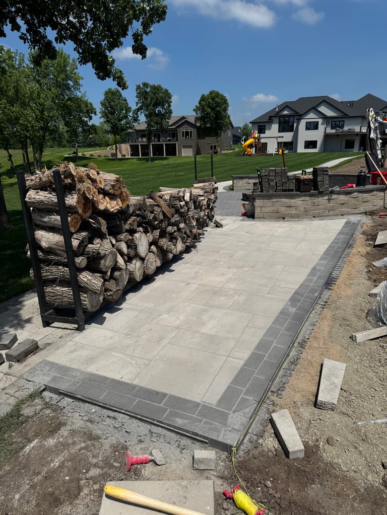 Suburban backyard patio under construction with stacked logs, concrete pavers, and residential homes visible in background
