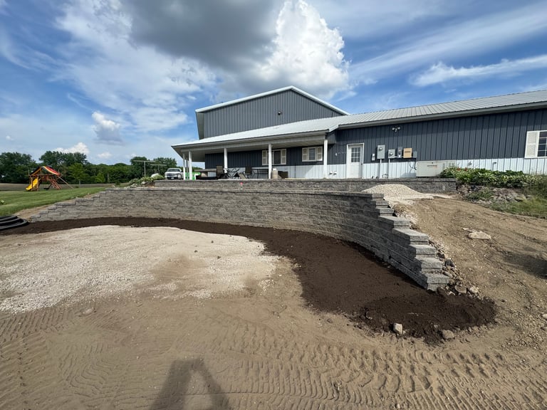 Modern metal-sided barn with porch under construction, surrounded by dirt earthwork and landscaping with blue sky above
