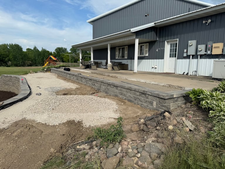 Construction site in front of a gray metal-sided barn with gravel, lumber, and equipment visible during renovation work
