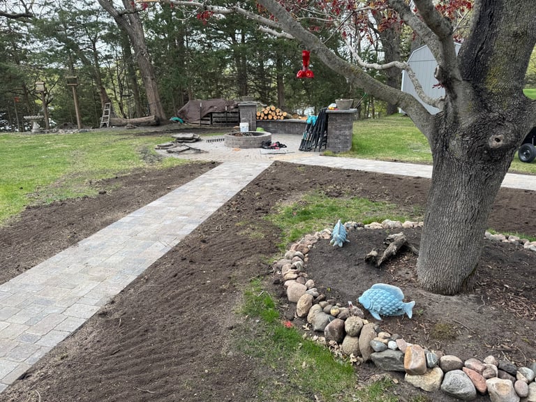 Park with stone-edged garden beds, paved pathways, and large shade tree in foreground. Picnic area and structures visible in background.