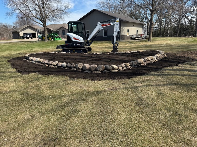 A small excavator with black and white coloring sits in the center of a circular stone border on a residential property with houses in the background