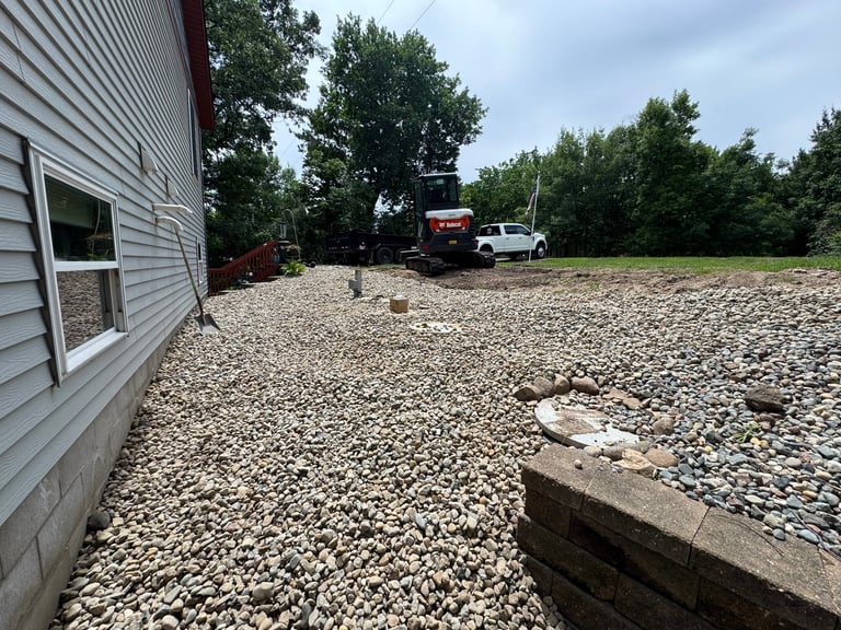 Gravel driveway beside a white house with vehicles and trees in the background under cloudy sky